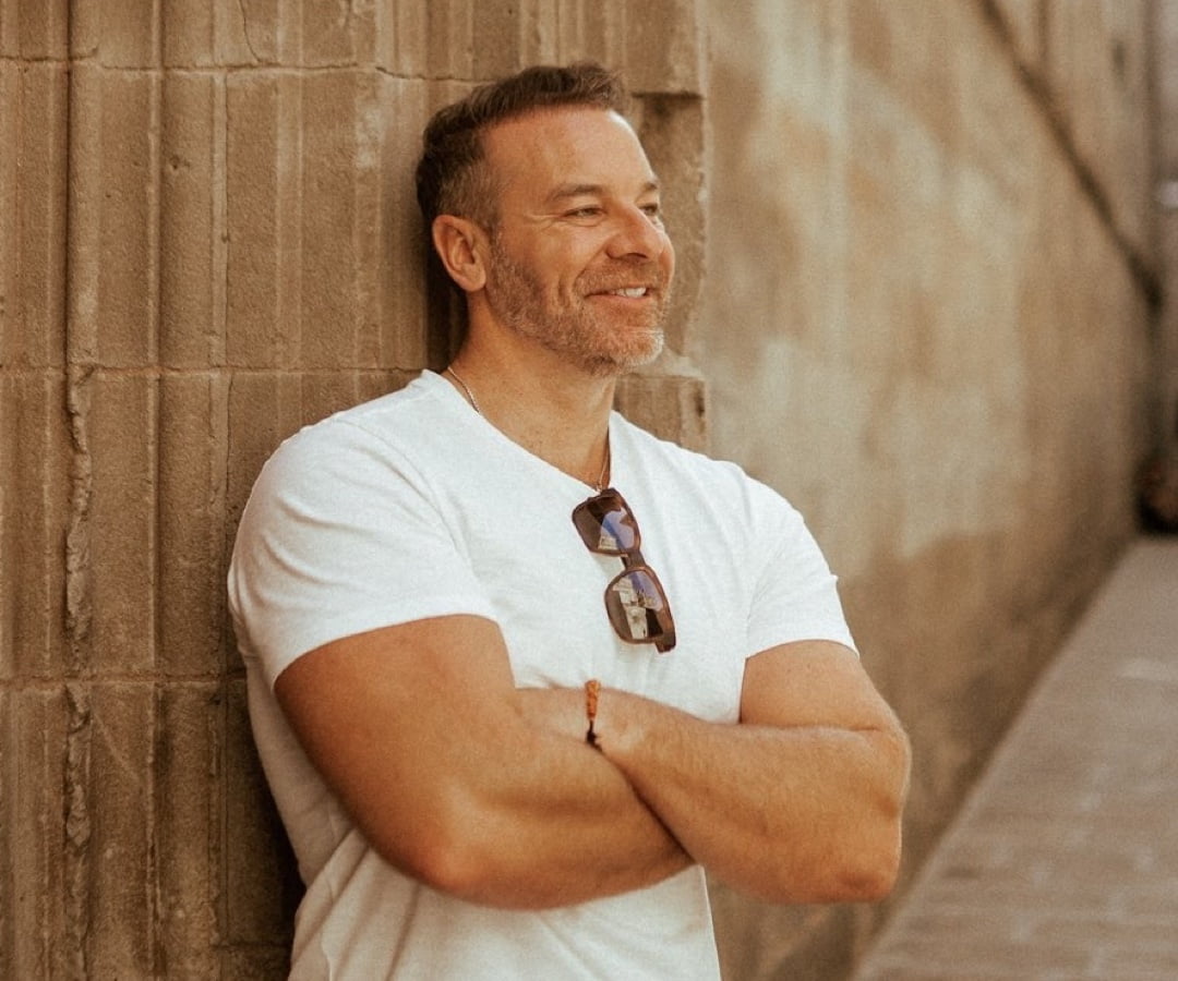 Man standing against a scenic coastal backdrop, arms crossed, wearing sunglasses, and smiling towards the camera.