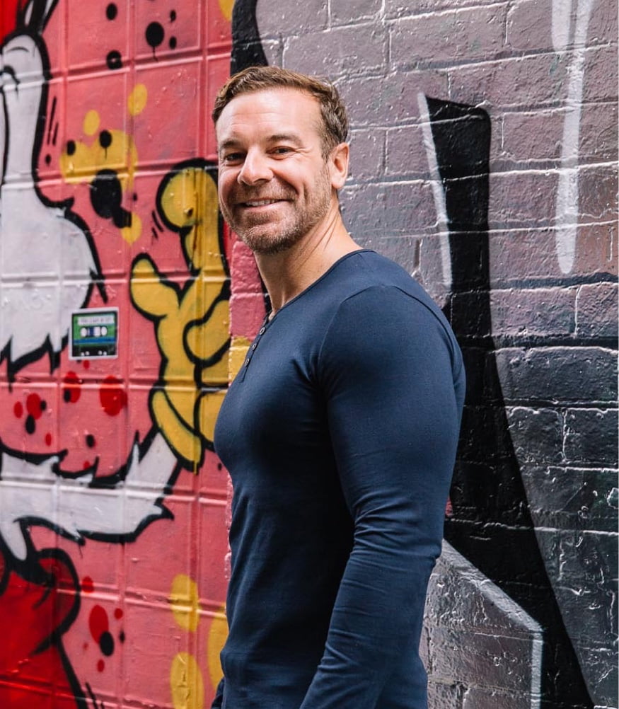 Man standing in front of a colorful street art mural, giving a side glance with a slight smile.