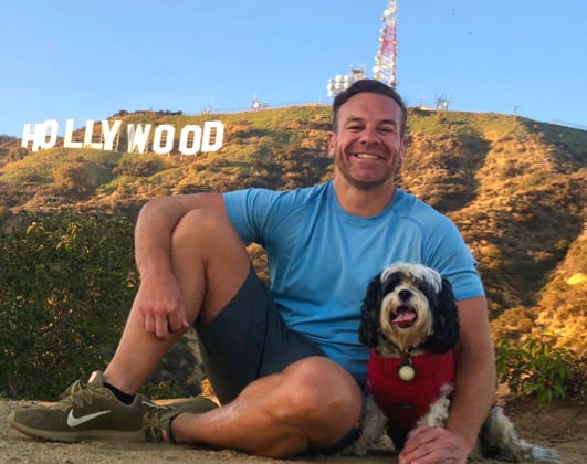 The man leaning against a tree with a blurred cityscape in the background, smiling at the camera, dressed in a gray t-shirt and jeans.