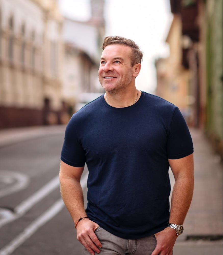 The man stands with crossed arms wearing a white polo shirt, in front of an ornate wooden door, with a charming smile.