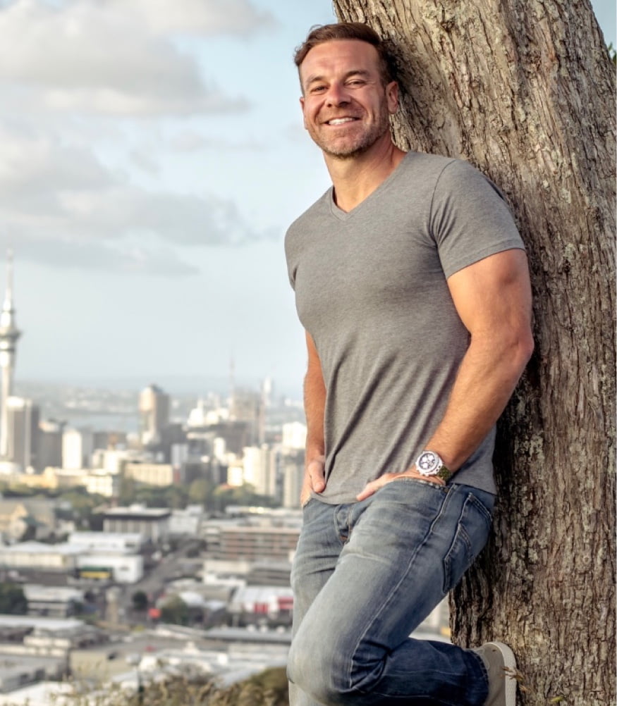 A portrait of the man in a blue long-sleeved shirt standing against a textured wall with graffiti, giving a pleasant smile.