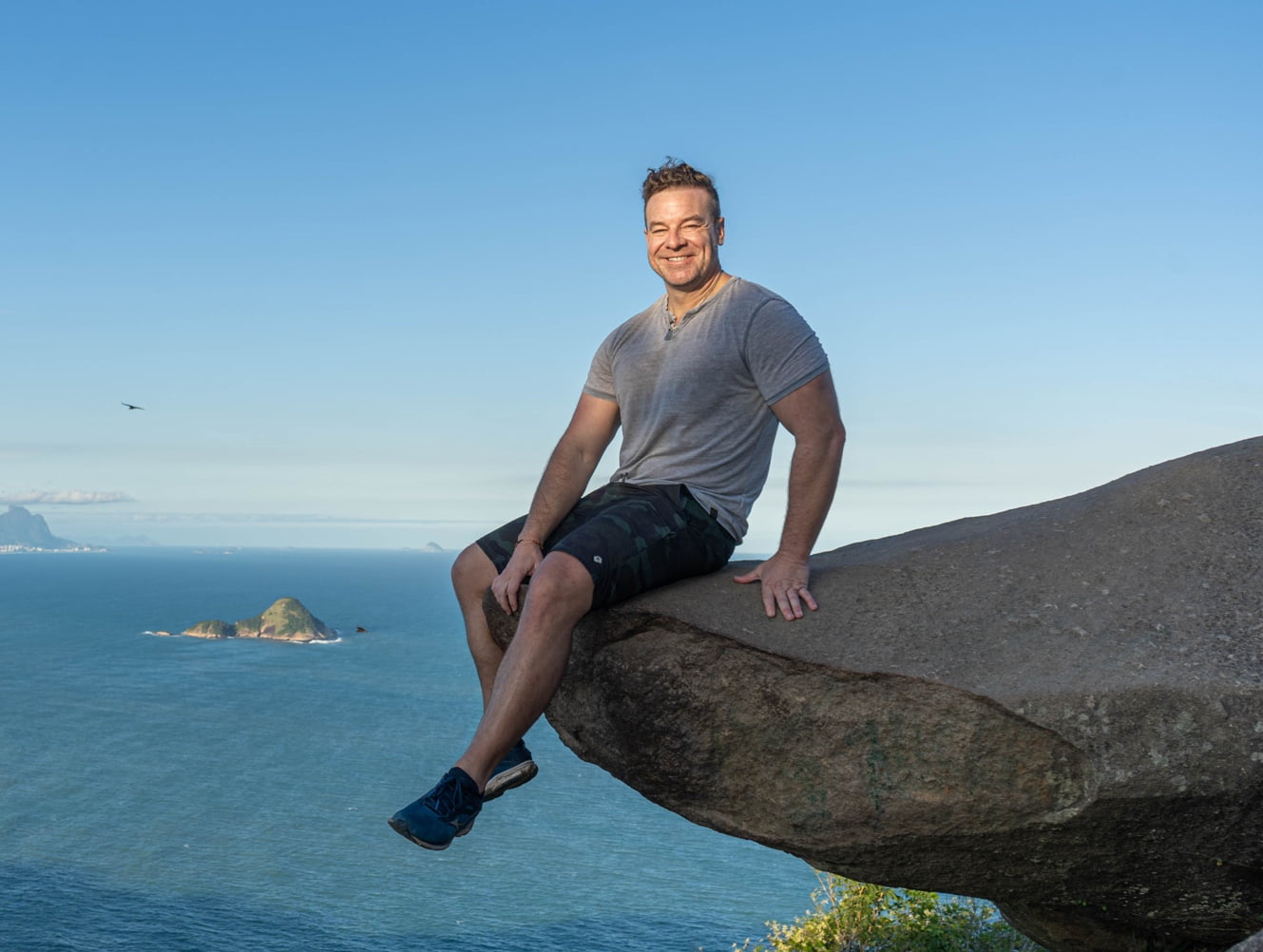 The man is sitting on the edge of a rock formation, with a vast ocean and islands in the background, sporting a relaxed and happy demeanor.
