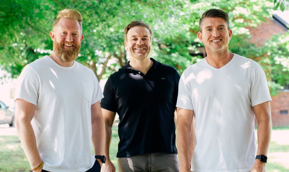 Three men smiling for a group photo outdoors, each dressed in casual attire, suggesting a friendly or team-oriented setting.